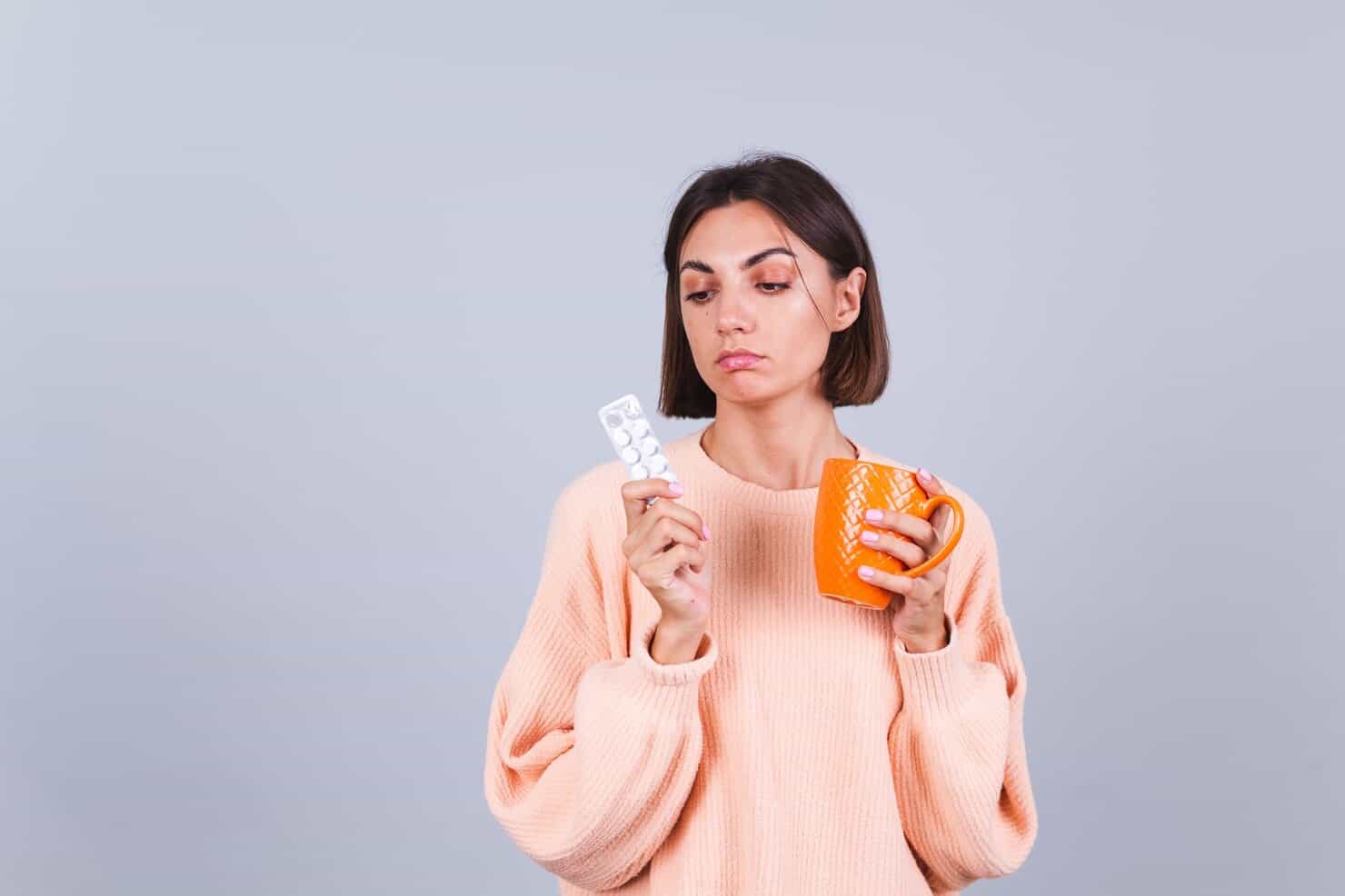 Woman in sweater on gray wall holds mug and pills